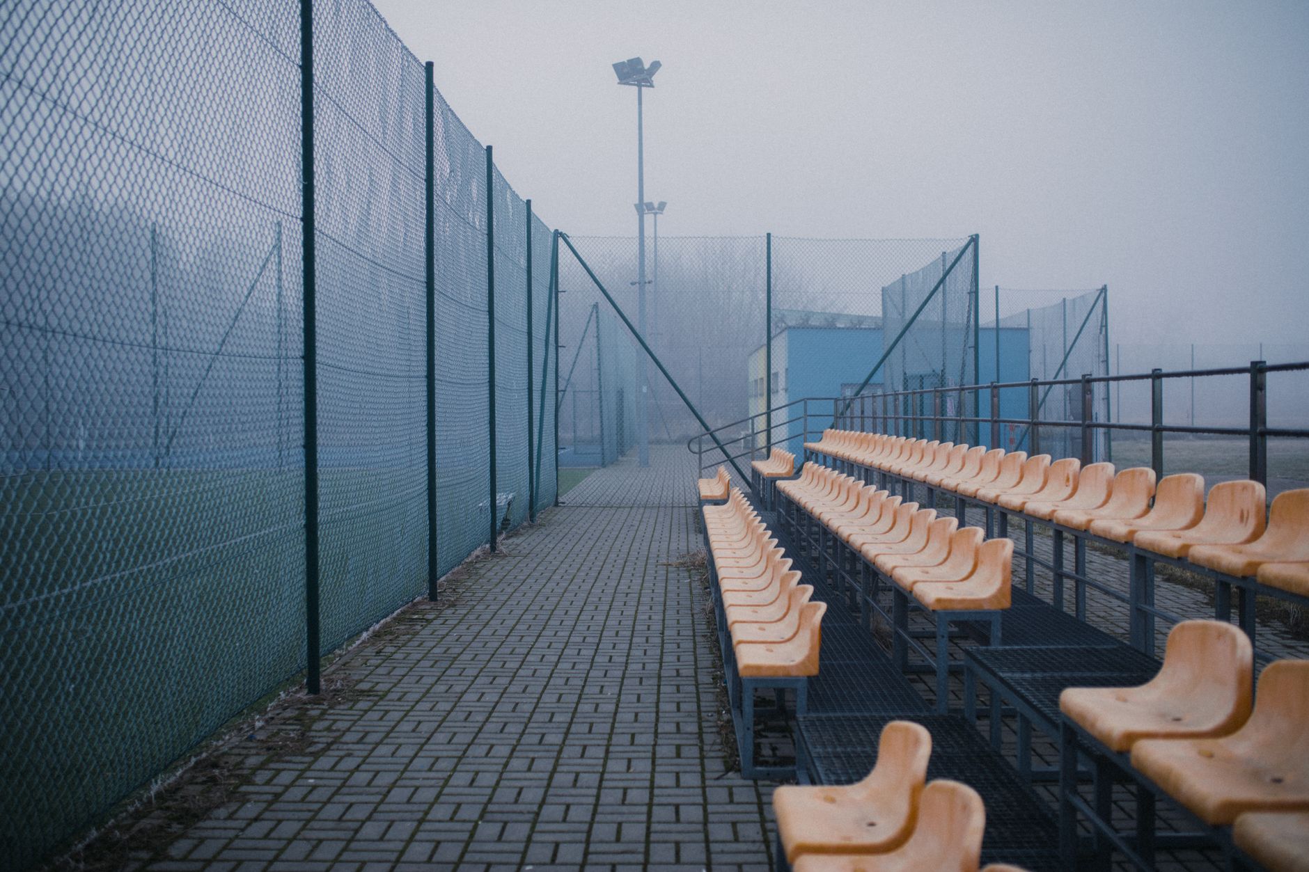 Tribune vide aux sièges orange au bord d'un terrain de football grillagé, dans la brume