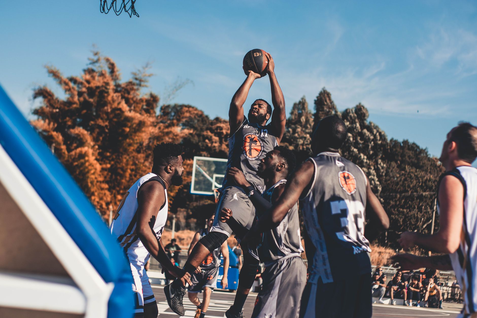 Un basketteur en maillot gris saute pour tirer, entouré de joueurs en gris et en blanc, sur un terrain extérieur sous un ciel bleu.